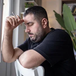 A man leaning on the back of a chair holding an unlit cigarette